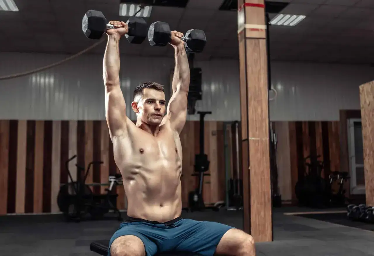 Man utför sittande hantelpress i ett gym. Han lyfter två hantlar över huvudet med raka armar.