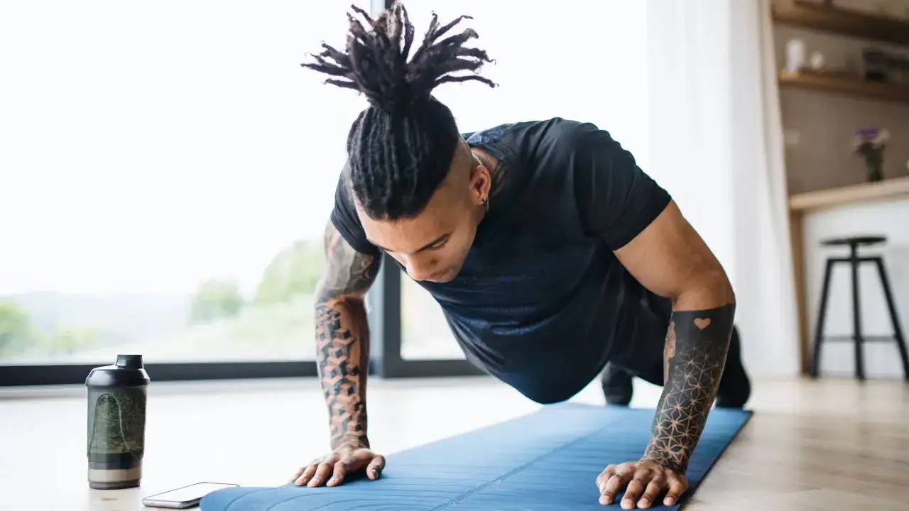 Man med dreadlocks gör armhävningar på en blå träningsmatta. Vattenflaska och mobil bredvid. Ett intensivt hiit pass hemma.