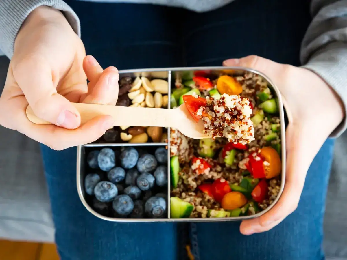 En person håller en matlåda med quinoa, grönsaker, nötter och blåbär. Perfekt för ett hälsosamt mellanmål viktnedgång.