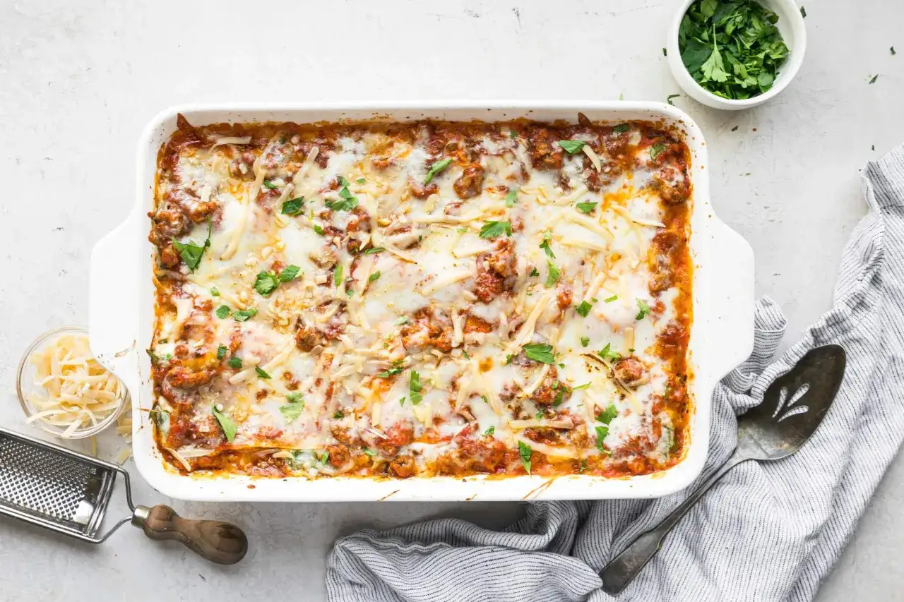 En gyllene, bubblande nyttig lasagne i en vit form, toppad med sm&auml;lt ost och f&auml;rsk persilja. Serveringssked och rivj&auml;rn bredvid.