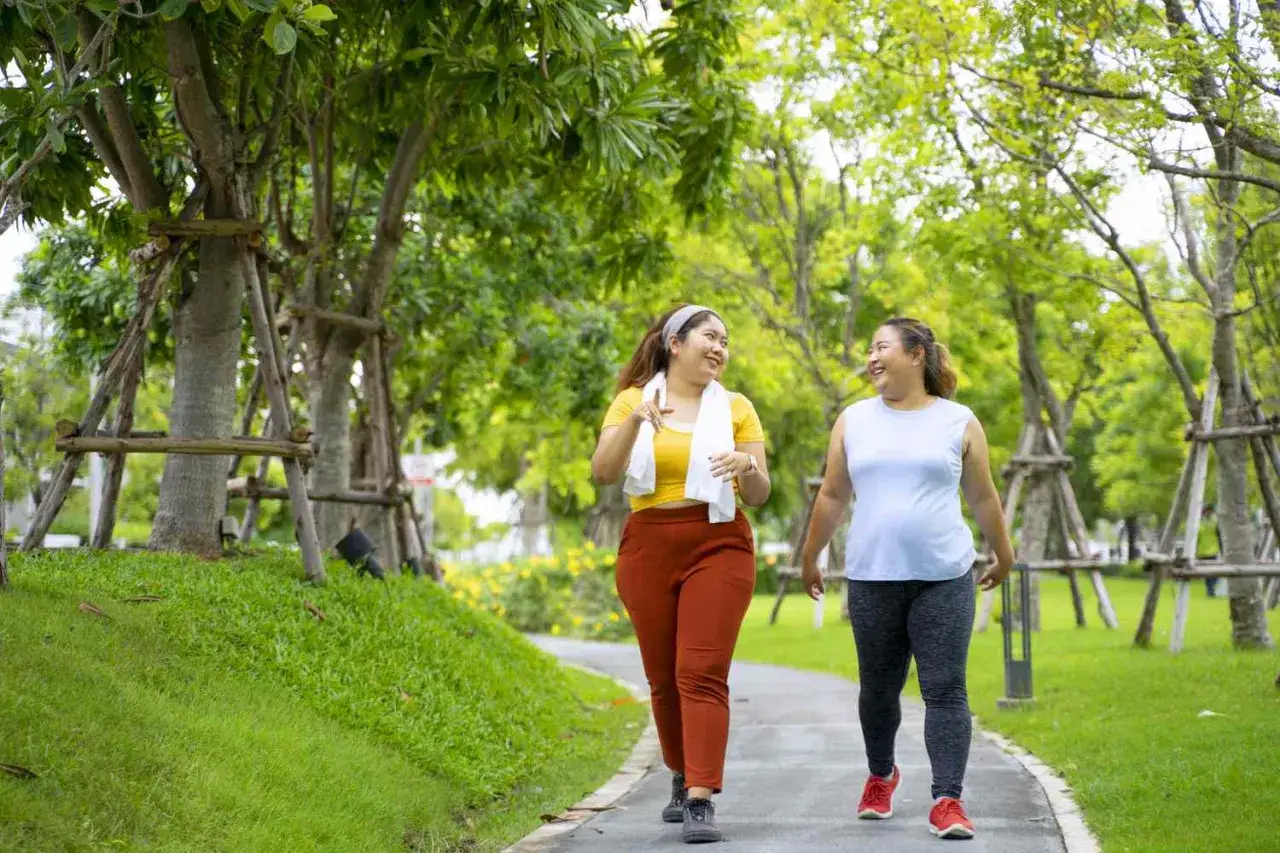 Två glada kvinnor promenerar i en park. De pratar och skrattar, kanske om hur lång tid det tar att gå 10000 steg.