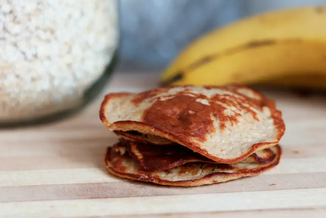 En hög med små, gyllenbruna pannkakor, perfekta som kalorisnåla kakor. I bakgrunden syns en burk med havregryn och en banan.