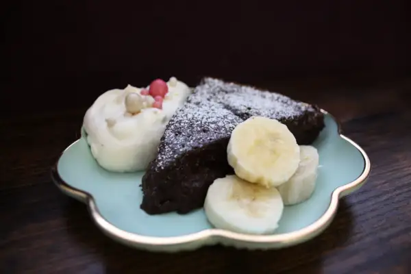 En bit nyttig kladdkaka med vispgrädde, bananskivor och strössel på en ljusblå tallrik.