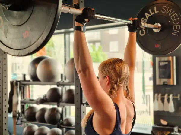 En kvinna lyfter en skivstång med vikter. Hon har flätor och träningshandskar. Frågan "hur blir man stark" besvaras med dedikation och hårt arbete på gymmet.