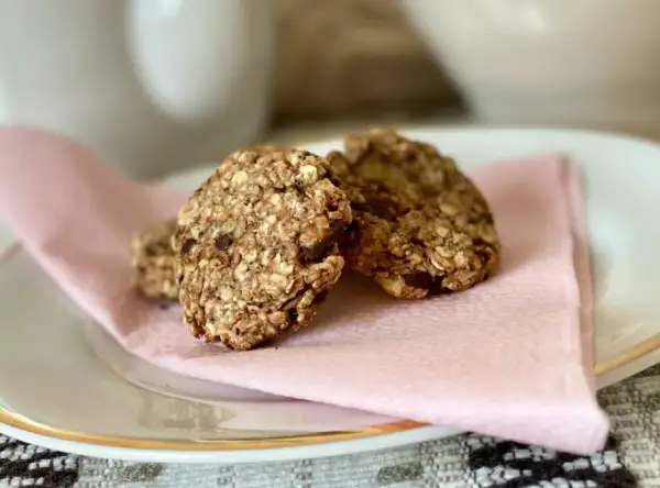 Tre hembakade, kalorisnåla kakor med havre och chokladbitar ligger på en ljusrosa servett på en vit tallrik.