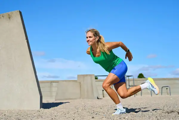 Kvinna i grönt linne och blå shorts utför ett intensivt hiit pass på en sandig yta, med betongkonstruktioner i bakgrunden.