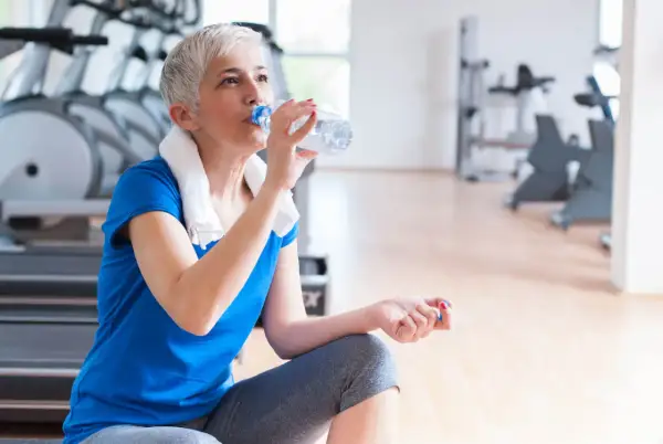 En kvinna med grått hår dricker ljummet vatten ur en flaska efter träningen på gymmet.