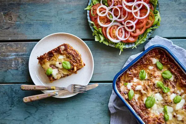 En bit nyttig lasagne på en tallrik bredvid en hel lasagneform och en fräsch tomatsallad med rödlök.